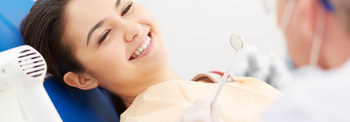 A woman sits in a dental chair smiling at a dentist who is holding a dental mirror.
