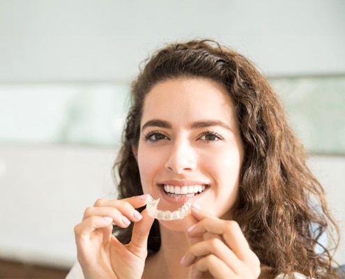 A woman with curly hair smiles while holding a clear dental aligner near her mouth, sitting in a brightly lit room.