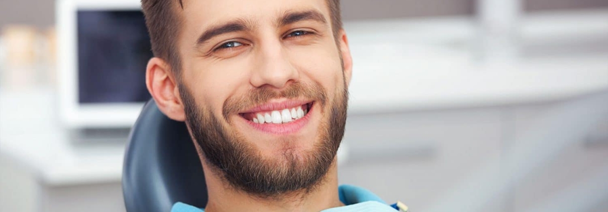 A man with a beard is sitting in a dental chair, smiling and showing his teeth. Dental equipment and a blurred background are visible.