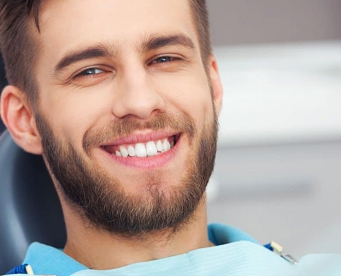 A man with a beard is sitting in a dental chair, smiling and showing his teeth. Dental equipment and a blurred background are visible.