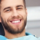 A man with a beard is sitting in a dental chair, smiling and showing his teeth. Dental equipment and a blurred background are visible.