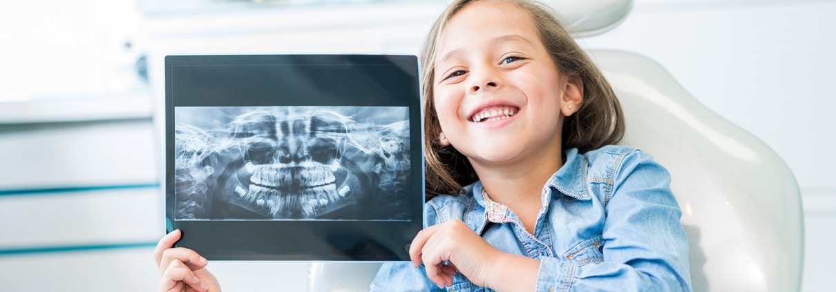 A young girl sitting in a dental chair smiles and holds up a dental X-ray image, showing her teeth and jaw.