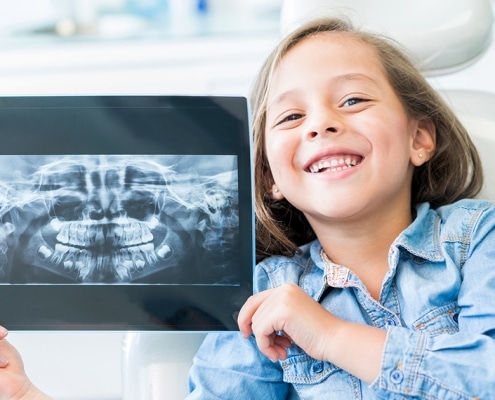 A young girl sitting in a dental chair smiles and holds up a dental X-ray image, showing her teeth and jaw.