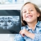 A young girl sitting in a dental chair smiles and holds up a dental X-ray image, showing her teeth and jaw.