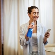Woman in pajamas stands in bathroom holding a water flosser and looking at her phone, smiling.