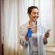 Woman in pajamas stands in bathroom holding a water flosser and looking at her phone, smiling.