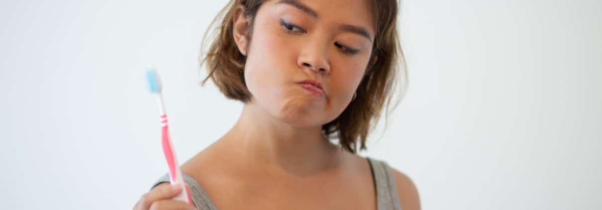 Young woman in a gray tank top looks skeptically at a toothbrush she is holding, standing against a plain white background.