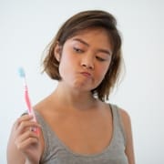 Young woman in a gray tank top looks skeptically at a toothbrush she is holding, standing against a plain white background.