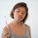 Young woman in a gray tank top looks skeptically at a toothbrush she is holding, standing against a plain white background.