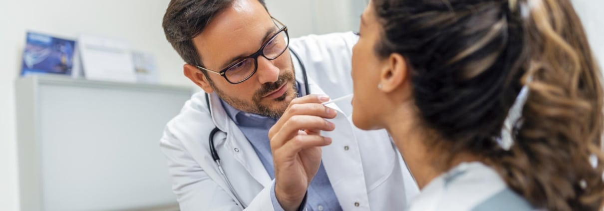 A doctor wearing a white coat examines a patients throat with a tongue depressor in a medical office.