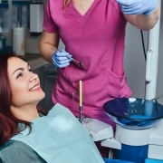 A woman sits in a dental chair smiling, wearing a bib, while a dental professional in pink scrubs and gloves prepares dental instruments nearby.