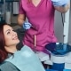 A woman sits in a dental chair smiling, wearing a bib, while a dental professional in pink scrubs and gloves prepares dental instruments nearby.
