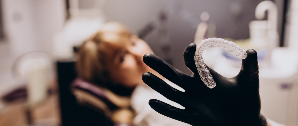 A gloved hand holds a clear dental aligner in focus, while a person sits blurred in the background on a dental chair.