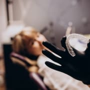 A gloved hand holds a clear dental aligner in focus, while a person sits blurred in the background on a dental chair.