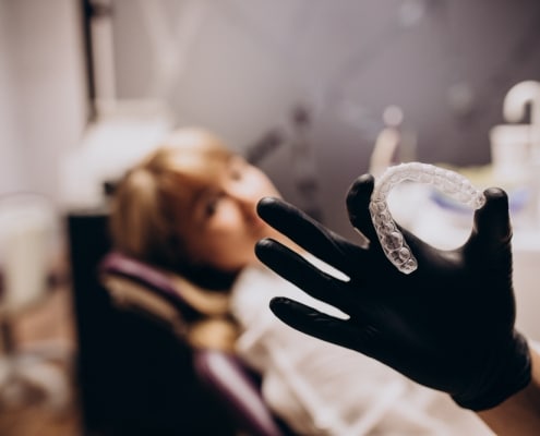 A gloved hand holds a clear dental aligner in focus, while a person sits blurred in the background on a dental chair.