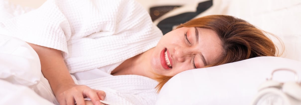 A woman lying in bed in a white robe appears to be in pain, clutching her stomach. An alarm clock is visible on the bedside table.