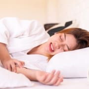 A woman lying in bed in a white robe appears to be in pain, clutching her stomach. An alarm clock is visible on the bedside table.