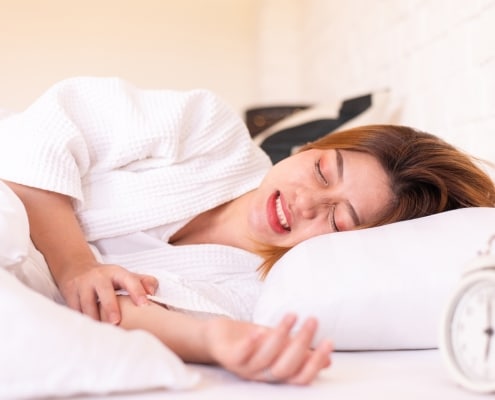 A woman lying in bed in a white robe appears to be in pain, clutching her stomach. An alarm clock is visible on the bedside table.