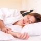 A woman lying in bed in a white robe appears to be in pain, clutching her stomach. An alarm clock is visible on the bedside table.